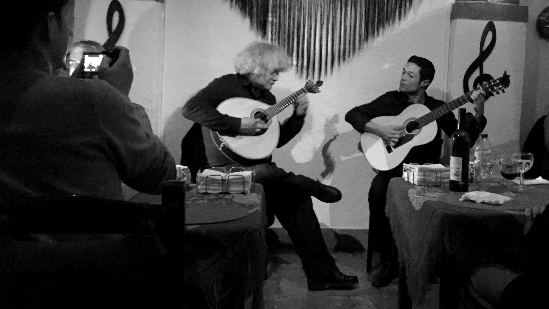 Two musicians, one with a pear-shaped Portuguese guitar and one with a classical guitar, play in a small whitewashed fado room in Alfama under a black fringed shawl, with diners at the tables around them