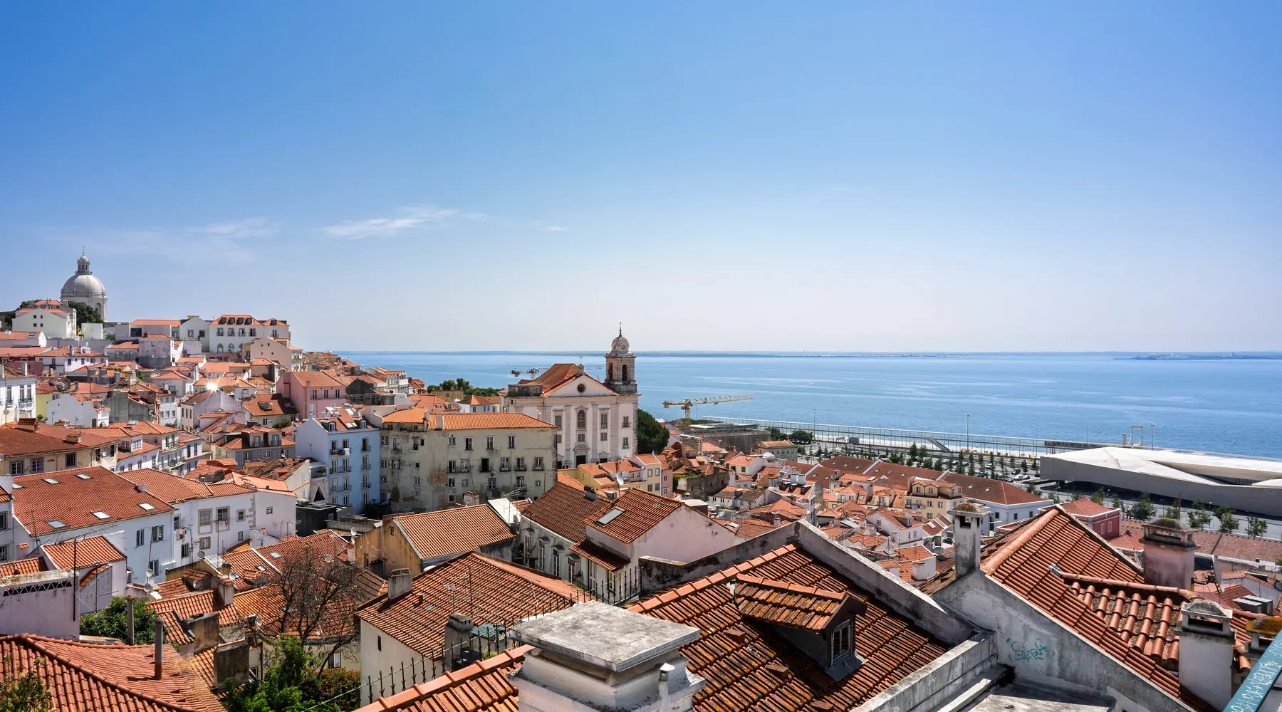 Terracotta rooftops of Alfama stepping down toward the Tagus river under a blue sky, with a church tower and the distant river