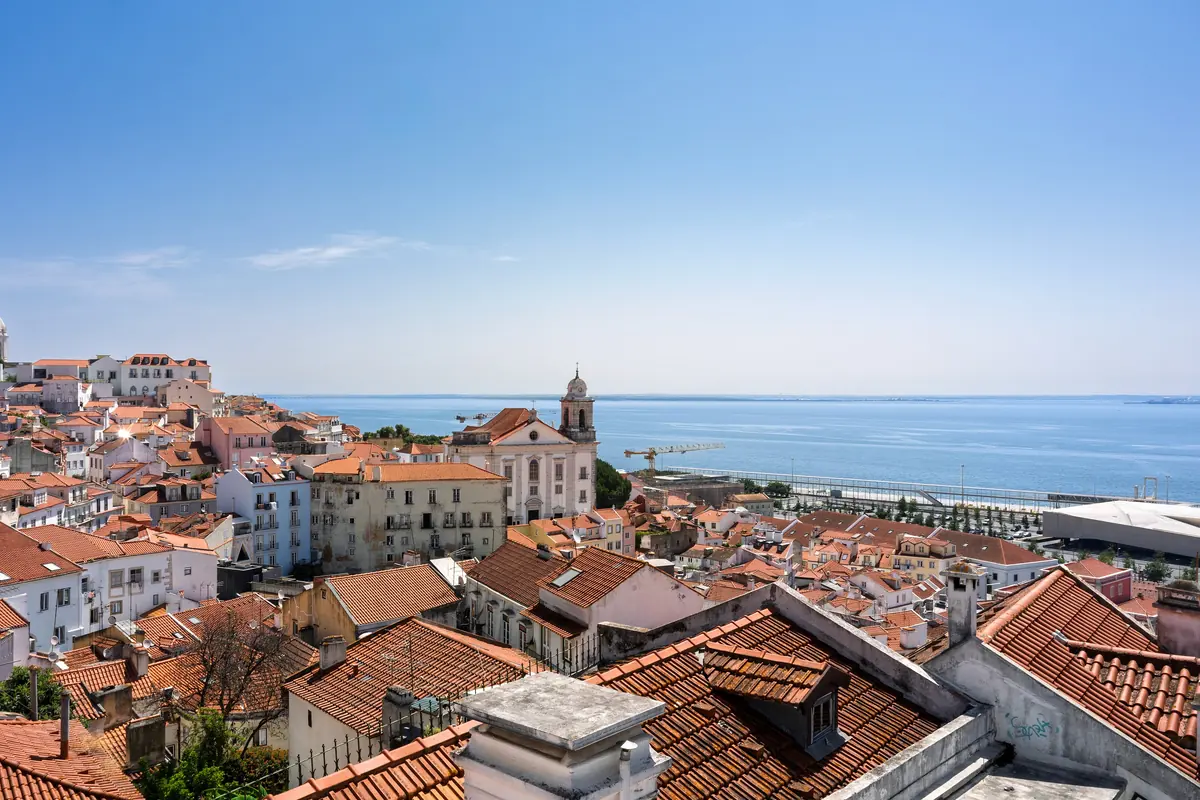Terracotta rooftops of Alfama stepping down toward the Tagus river under a clear blue sky, with a church tower and the river in the distance