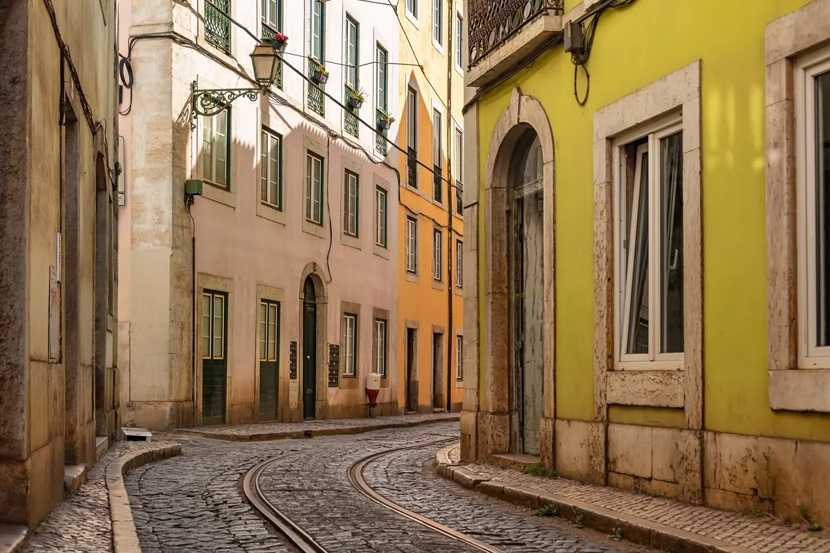 A curving cobbled lane in Alfama with tram rails set into the paving, between pale pink, yellow and ochre facades