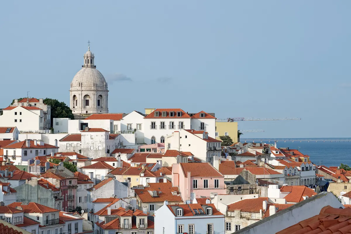 White and terracotta-roofed houses of Alfama tumbling down a hillside, with the white dome of the National Pantheon and the Tagus river behind