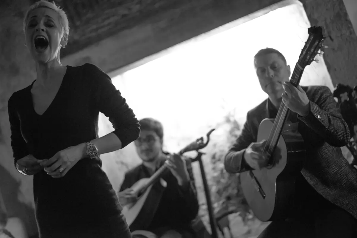 A woman in black sings with her mouth open wide while a guitarist plays a Portuguese guitar and another plays a classical guitar behind her, in black and white