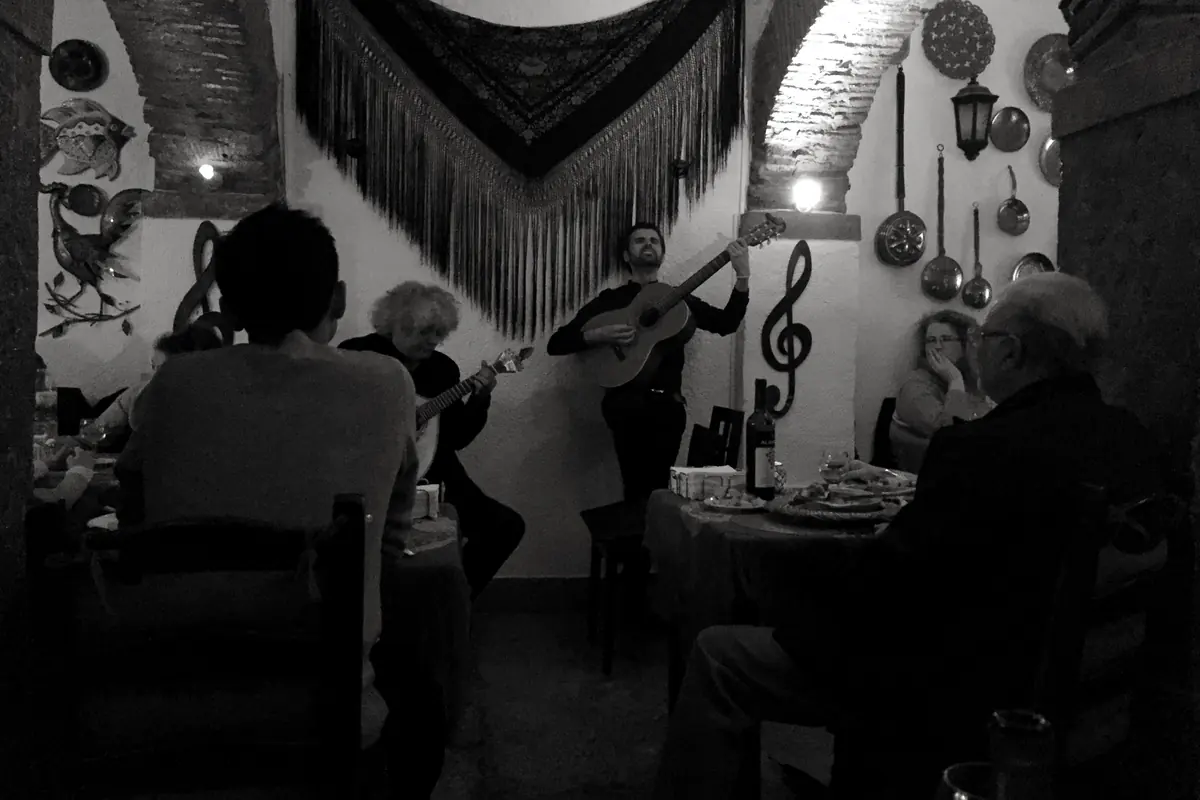 A singer and a guitarist perform in a small whitewashed fado room under a black fringed shawl, with diners at tables around them